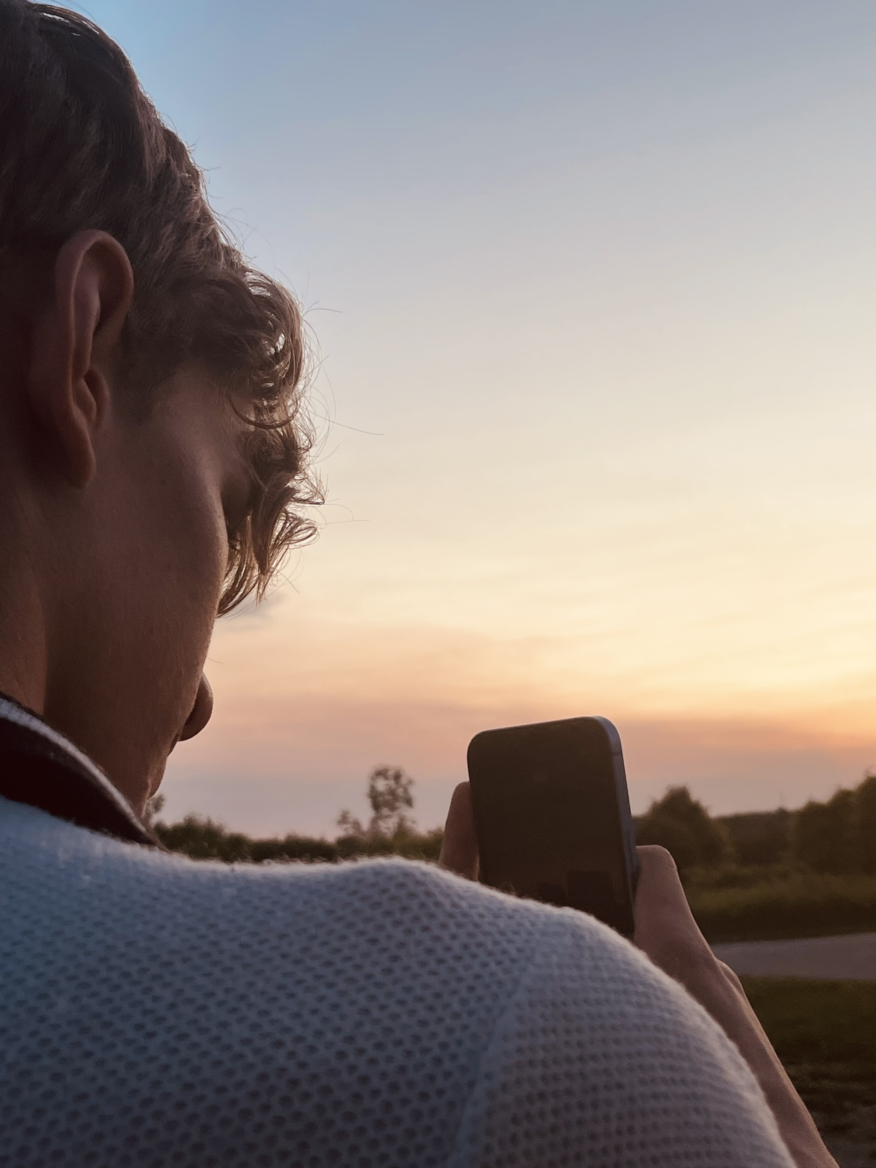 The shoulder of a blonde man taking a photo of the sunset in front of him.