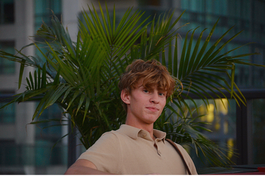 Blonde-haired man looking at the camera with a green bush behind him.