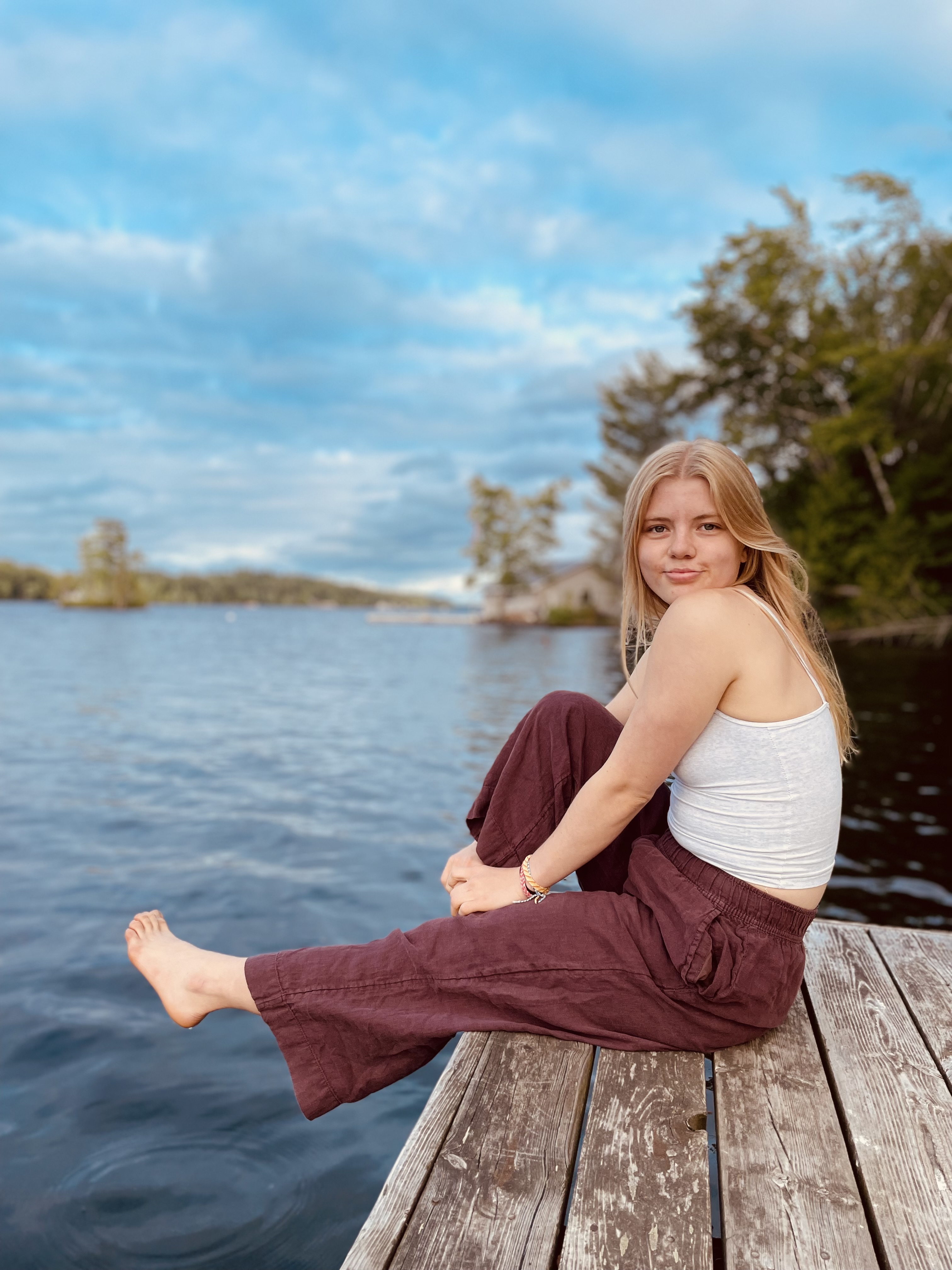 A blonde-haired girl sitting on a wooden dock next to a lake with her leg out, smiling at the camera.