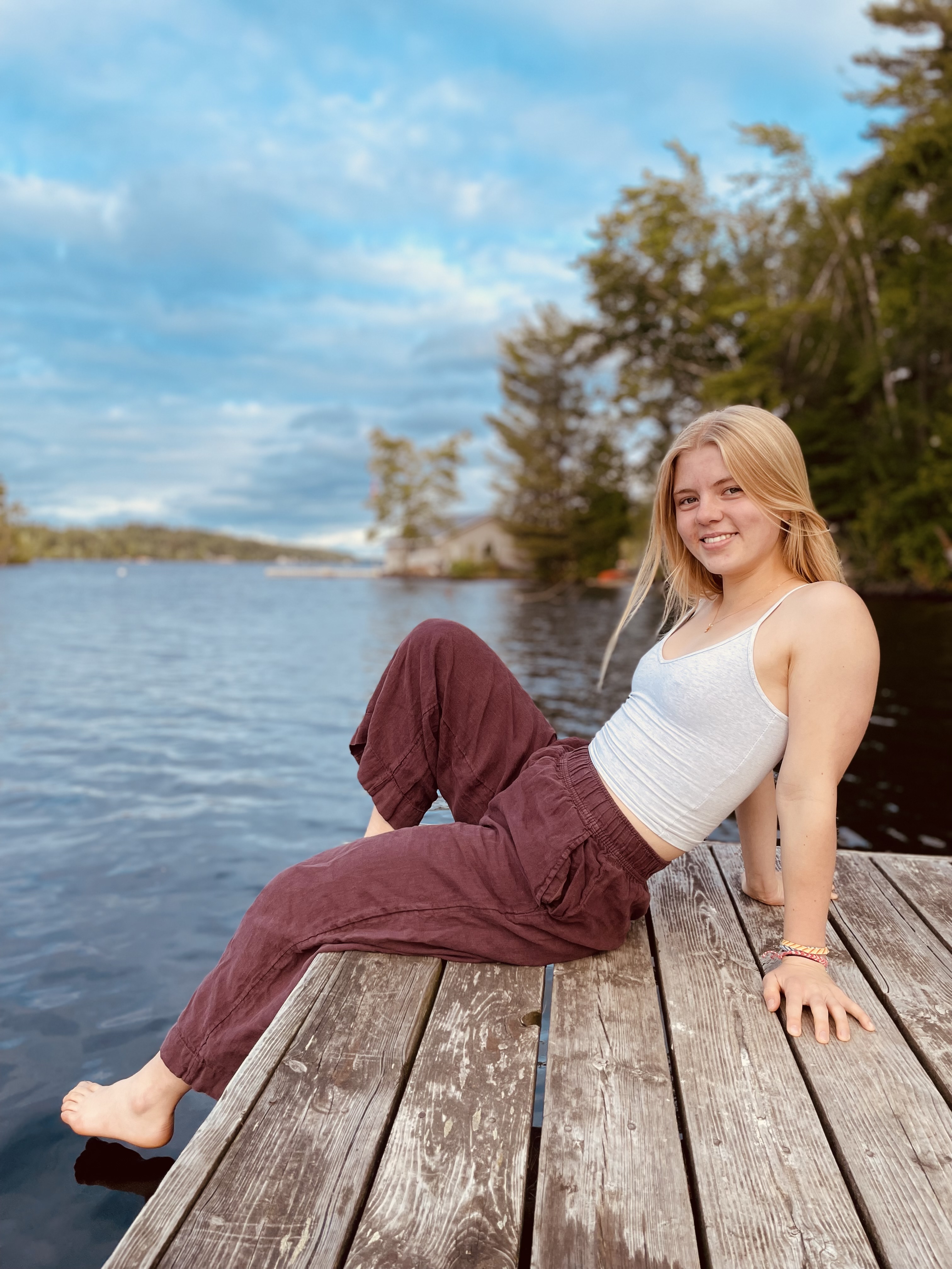 A blonde-haired girl sitting on a wooden dock next to a lake, smiling at the camera.