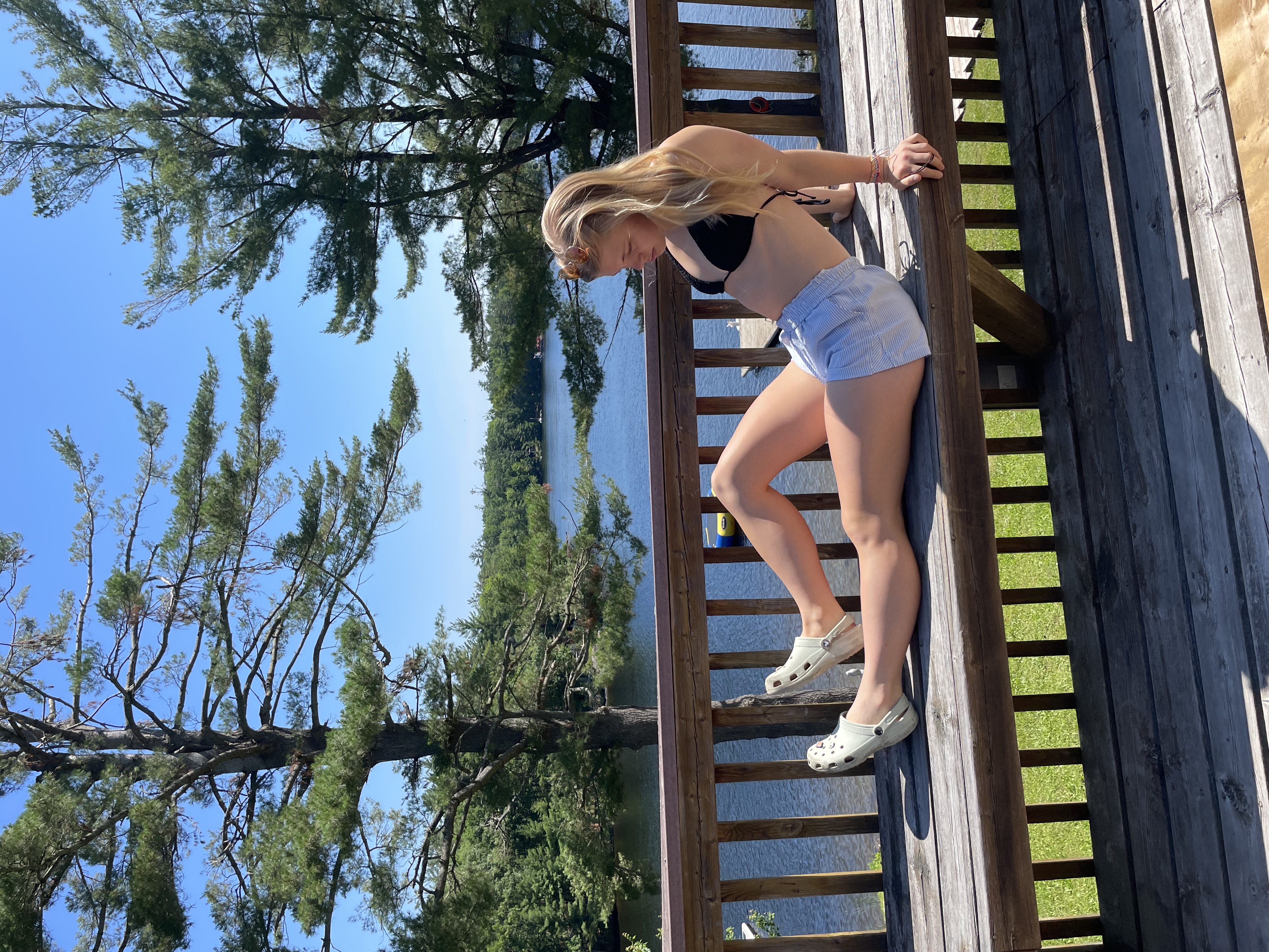 A blonde haired girl sitting on a wood bench with a green field and blue lake behind her. .