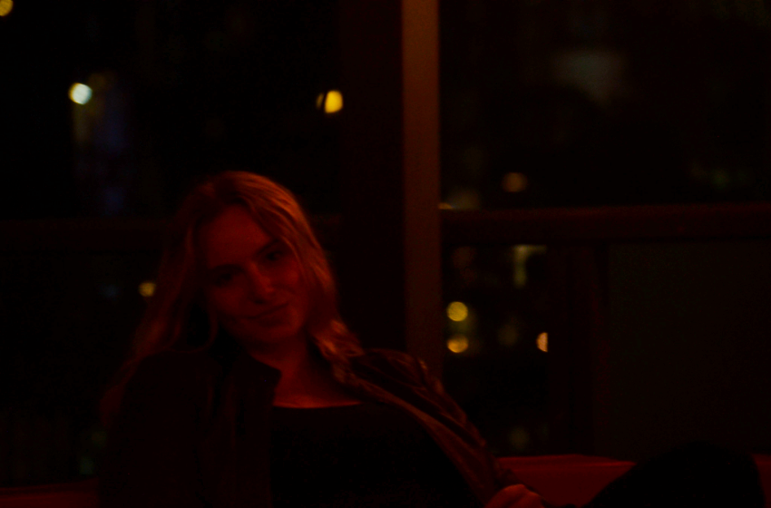 A blonde haired girl sitting in the dark, illuminated by red light.