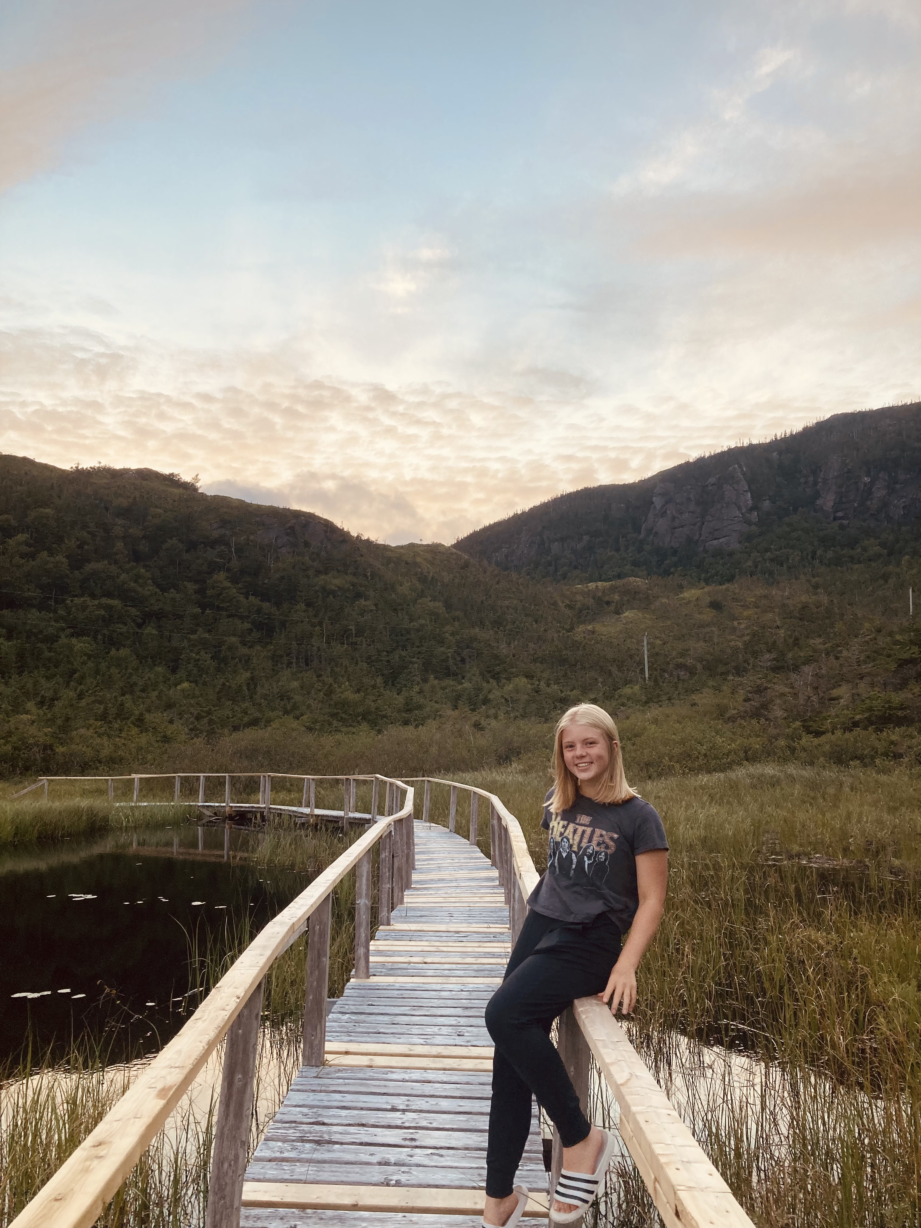 A blonde woman leaning on a railing next to a pond with mountains and a sunset behind her.