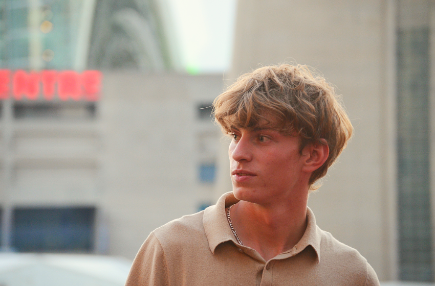 A blonde man looking to his side with a white building behind him.