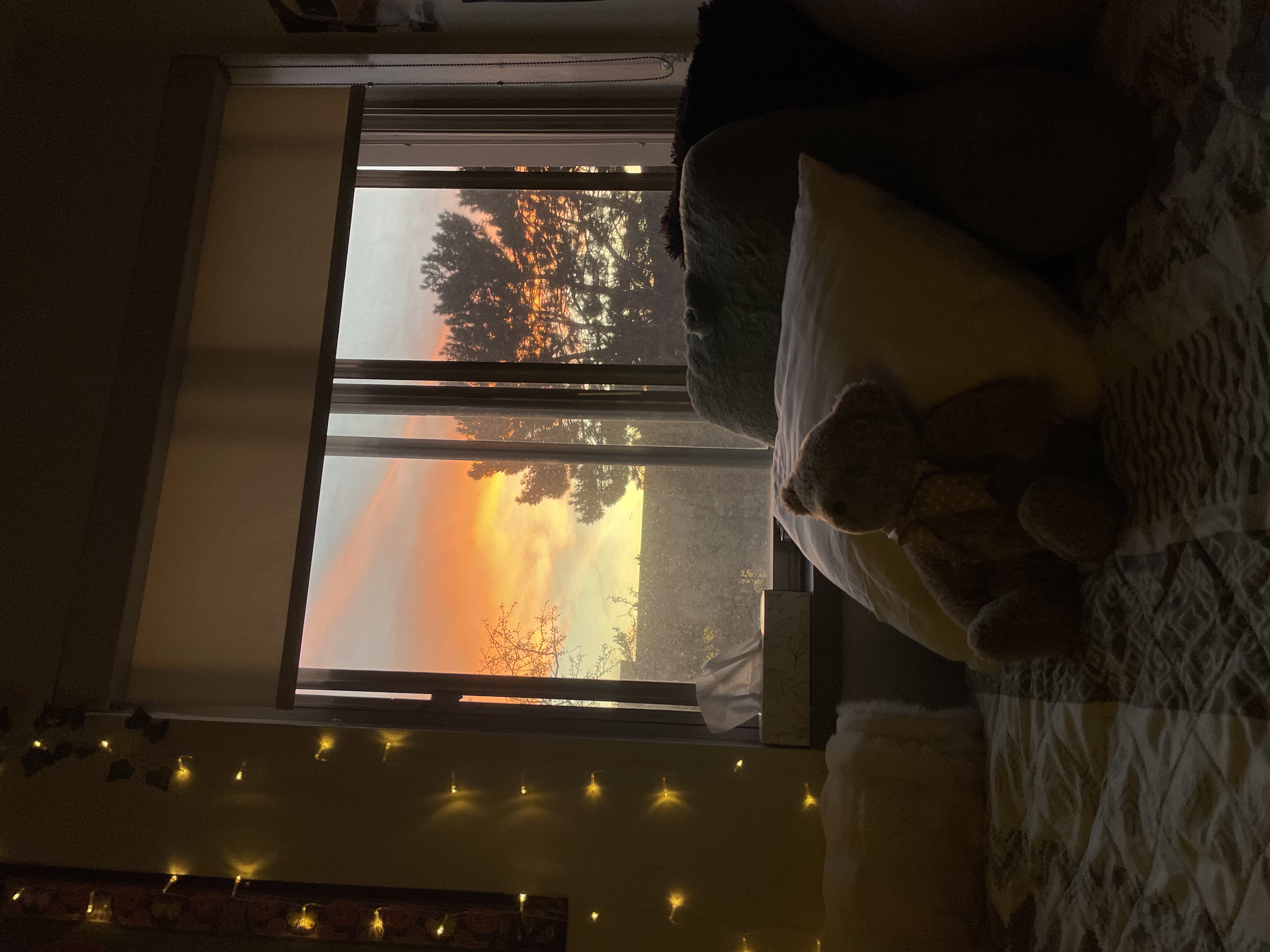 A colourful sunset outside of the window, seen behind a made bed in a dark room.