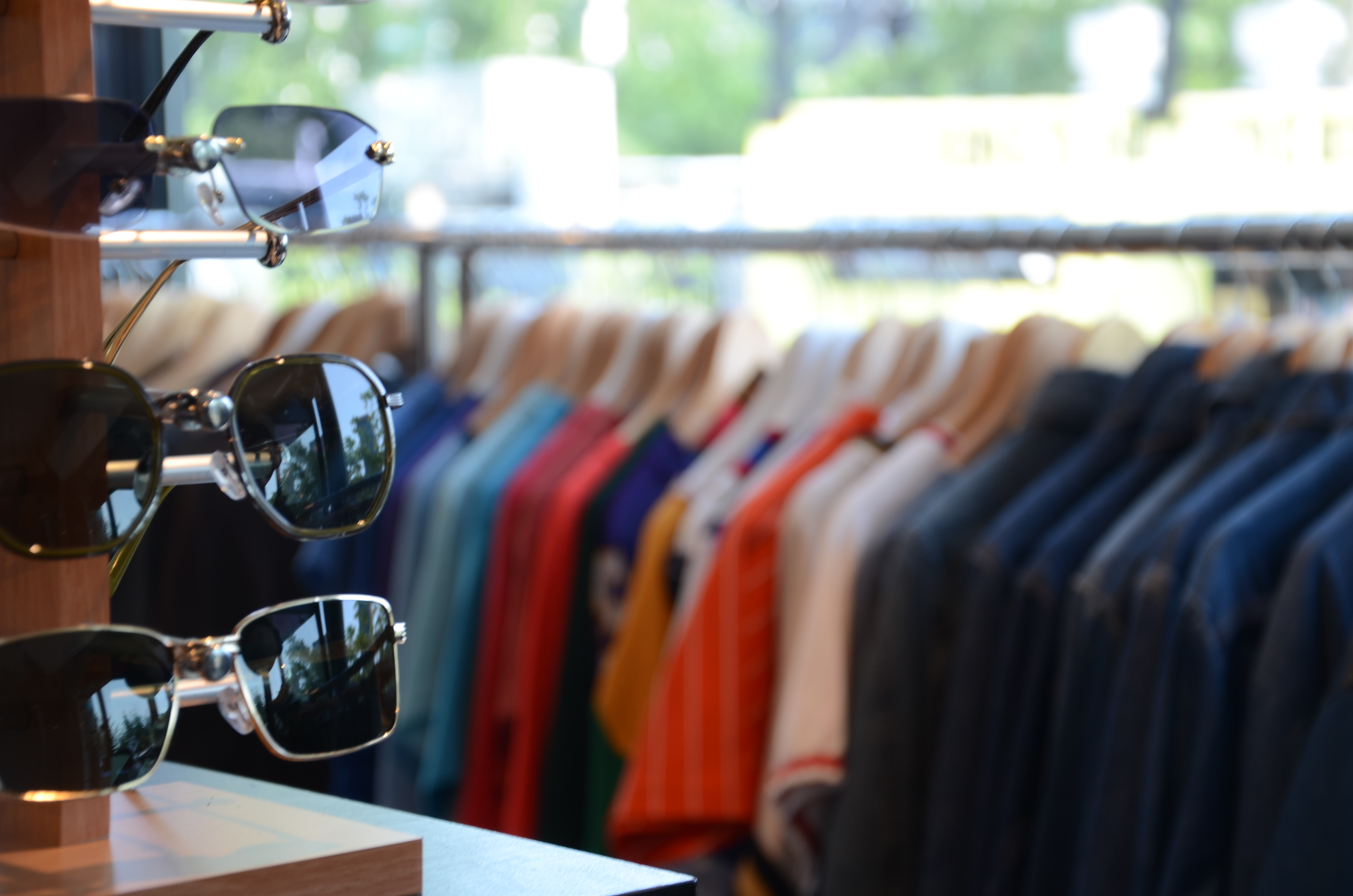 A sunglass rack in a thrift store with a rack of colourful clothes in the background.