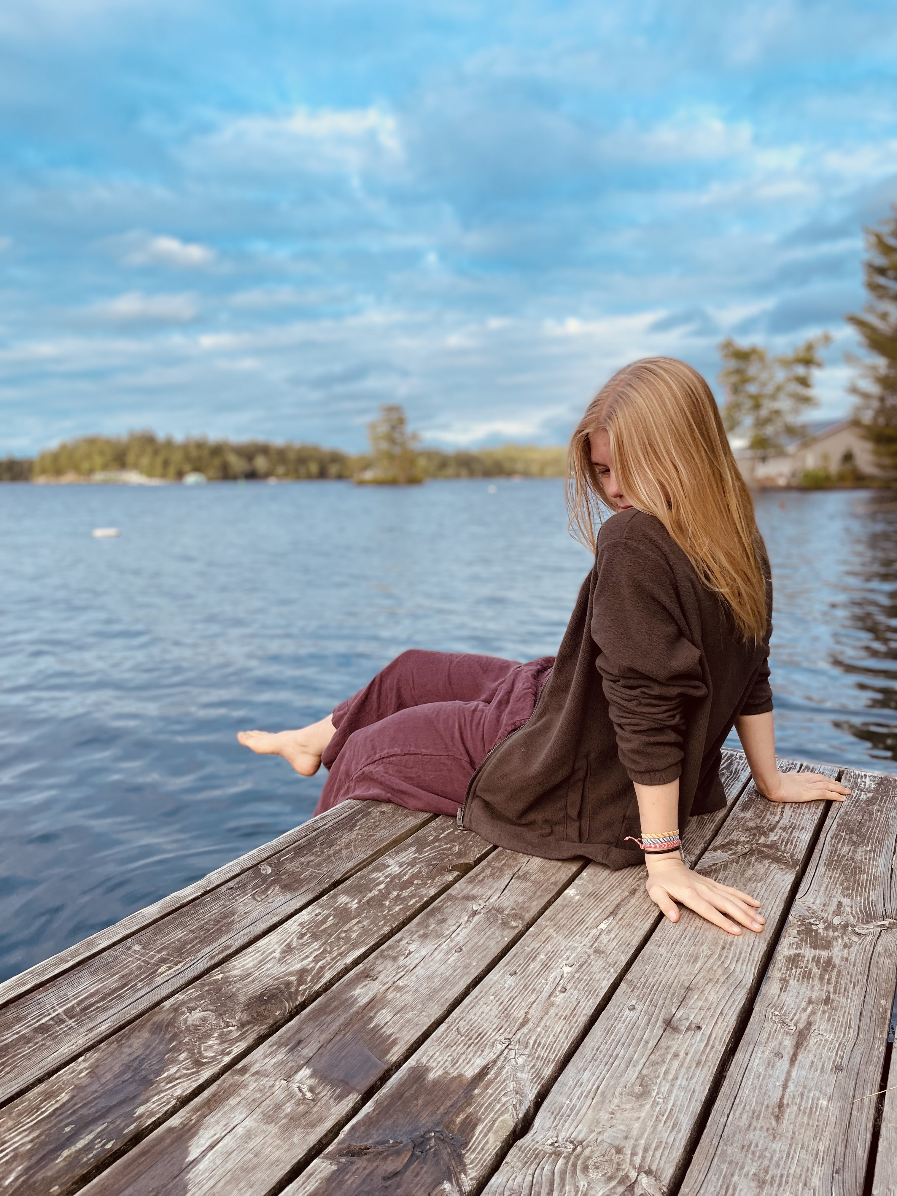A blonde-haired girl sitting on a wooden dock on next to a lake.