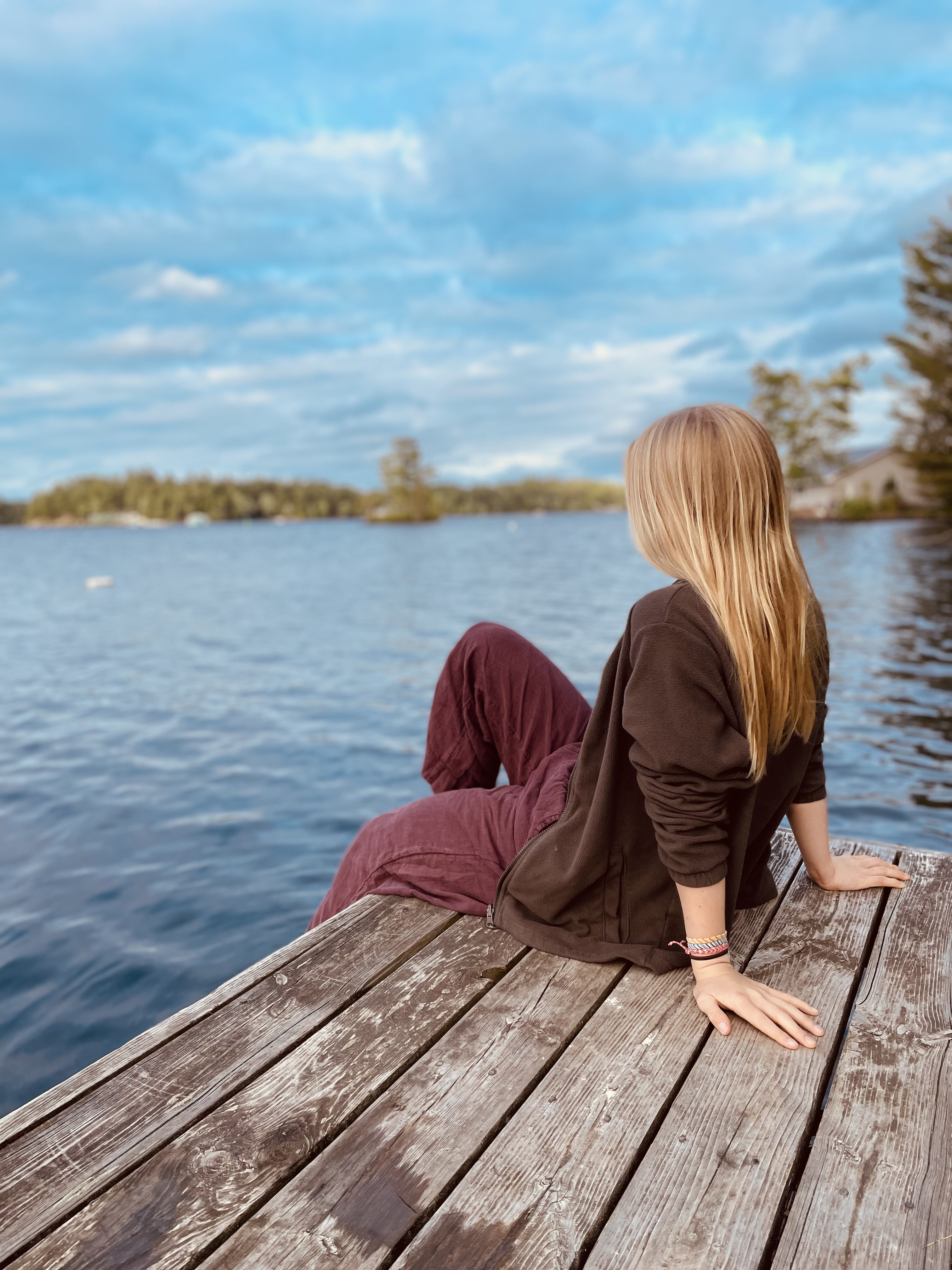 A blonde girl sitting on a wooden dock next to a lake.