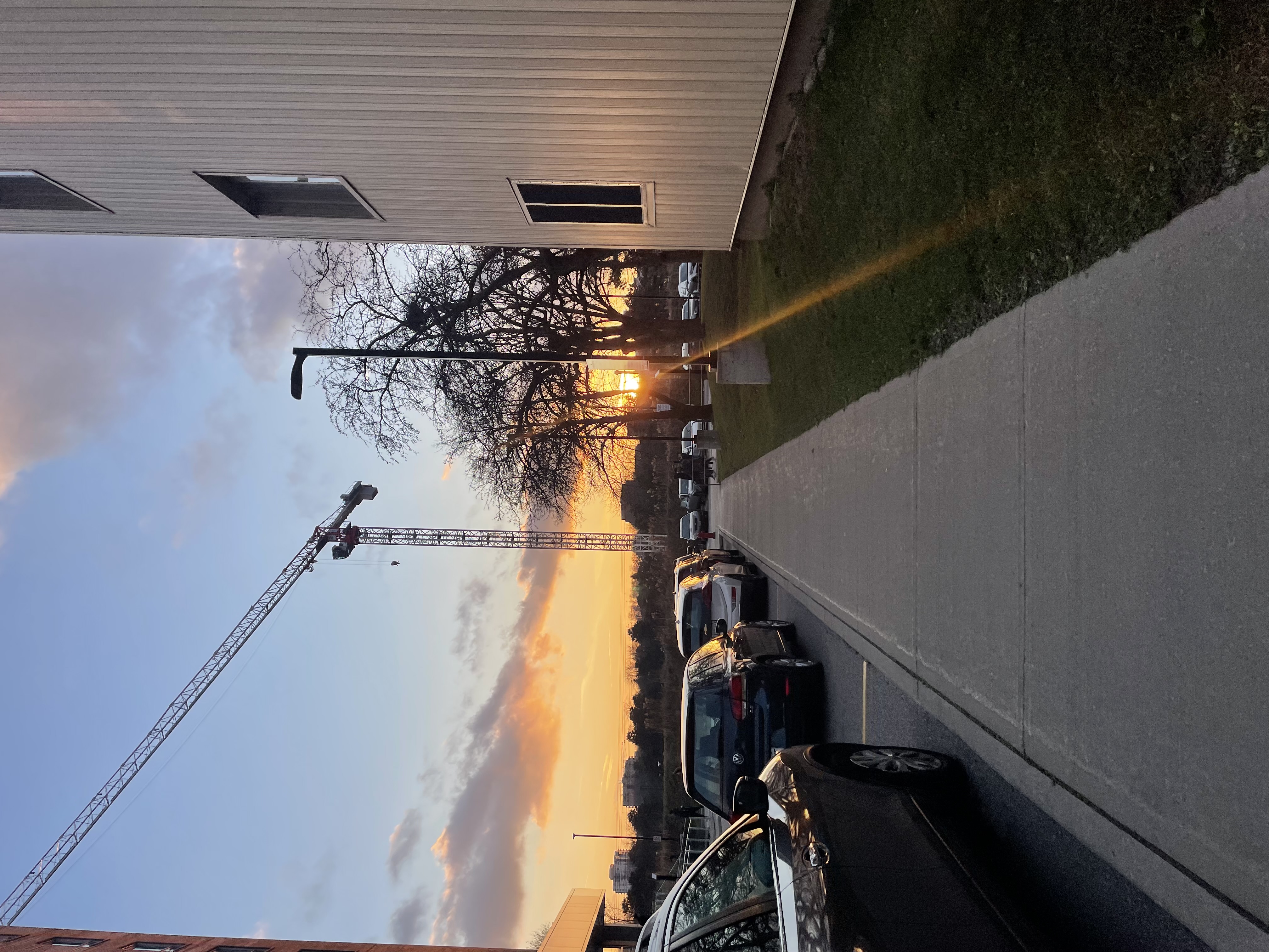 The sun setting behind a white building next to a line of parked cars.