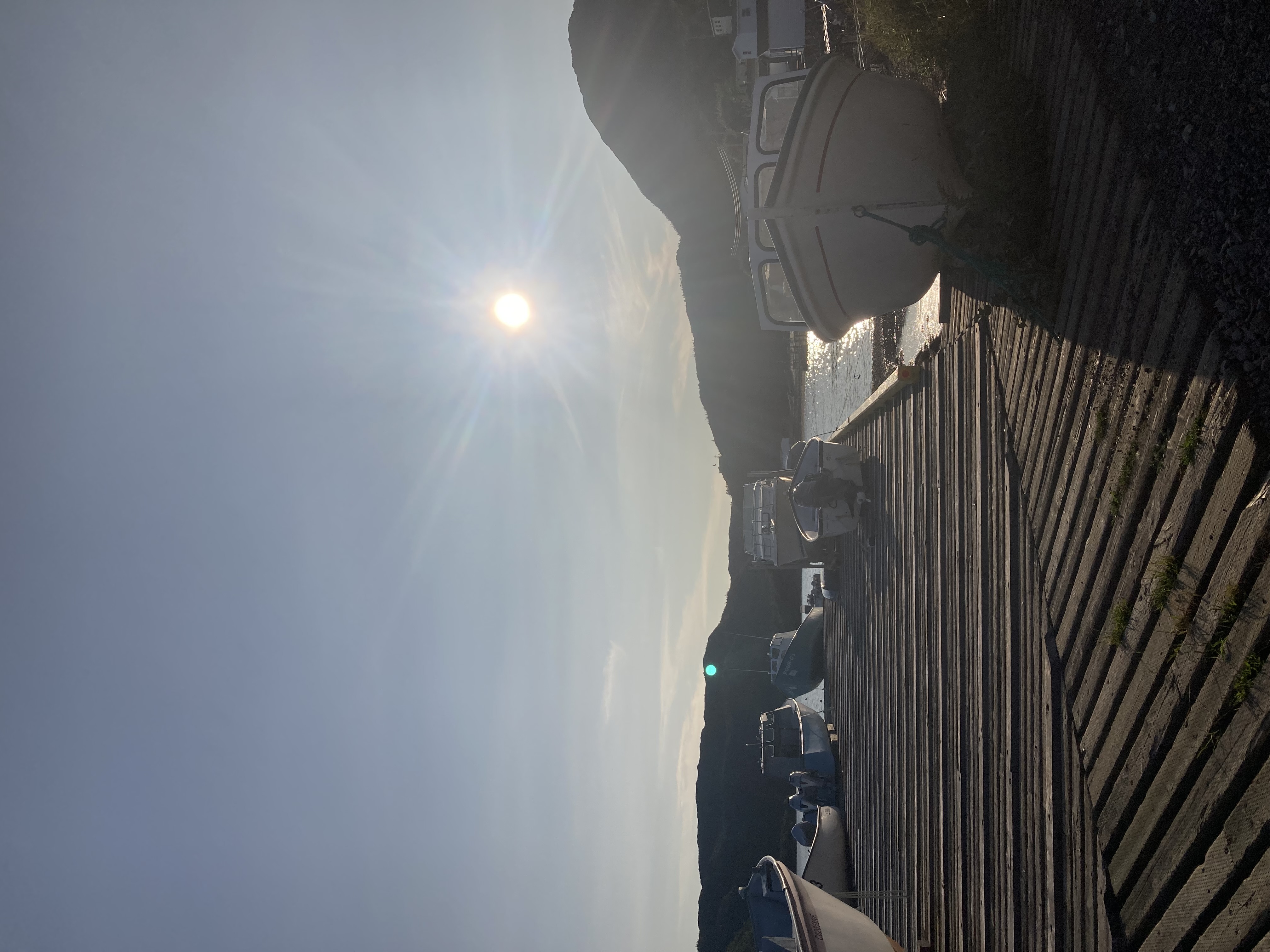 A set of white boats on a wooden slat pier next to the ocean, with the sun starting to set in the background.